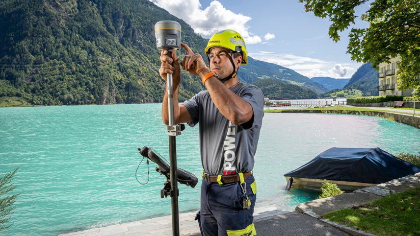 Technischer Spezialist misst am See mit GPS-Gerät; Berge und türkisfarbenes Wasser im Hintergrund.