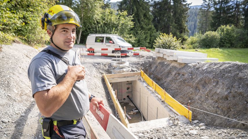 Netzelektriker mit Helm steht an einer Baustelle neben einem Kabelschacht; Absperrungen und Material im Hintergrund.