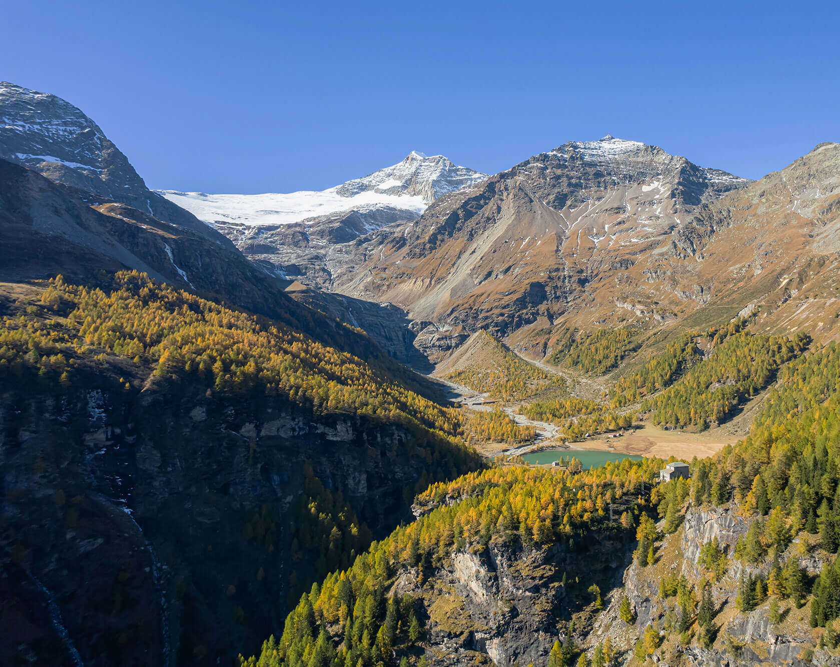 Blick über ein alpines Tal mit Gletscher, Lärchenwald und kleinem See in herbstlicher Berglandschaft.