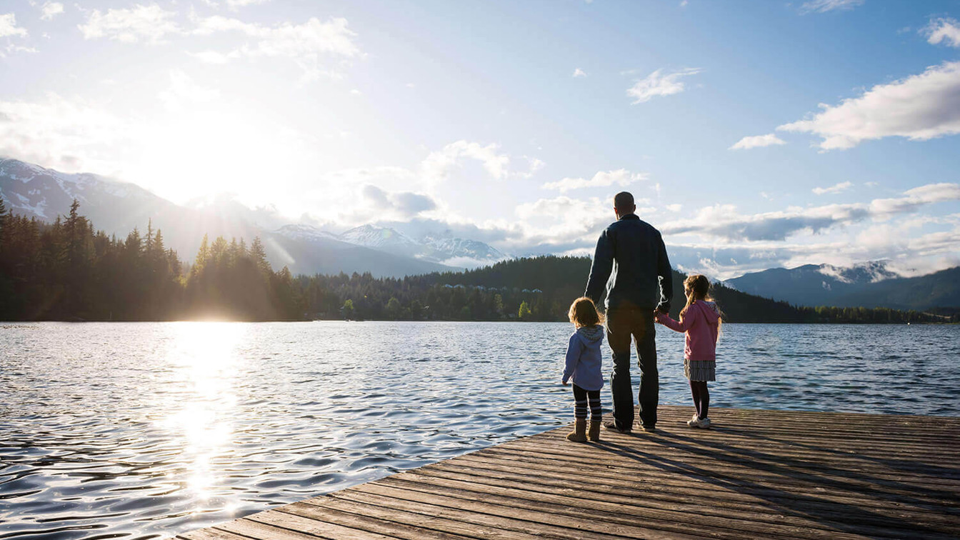Vater mit zwei Kindern steht auf einem Steg am See und blickt in die Berge bei Sonnenuntergang.